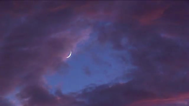 Tinted clouds sweep past a crescent moon at golden hour.