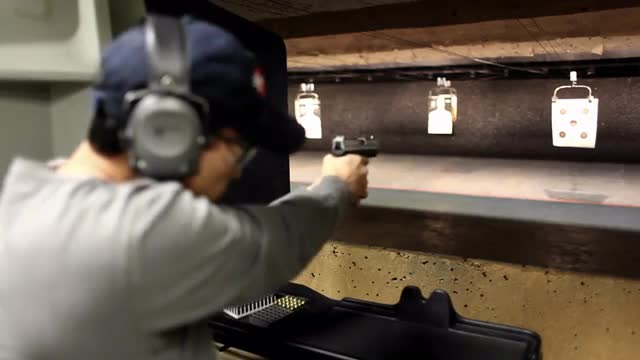 A man looks at the camera and then fires a hand gun at a target at an indoor shooting range.