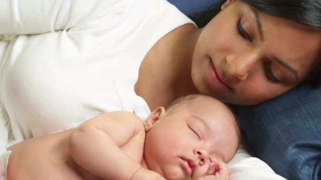 A beautiful woman lies on a couch with her newborn baby sleeping on her arm