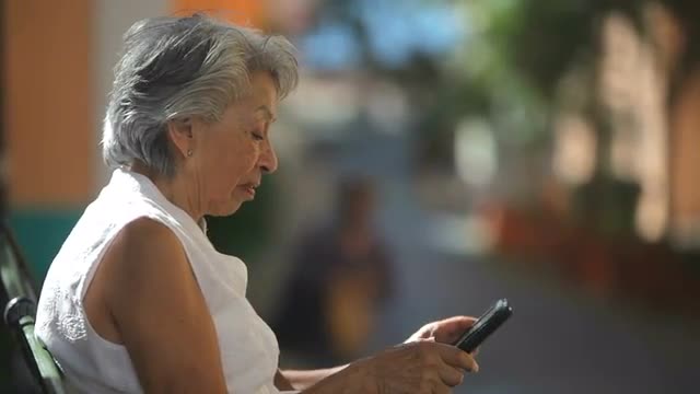 An old lady sits on a park bench and uses her tablet computer