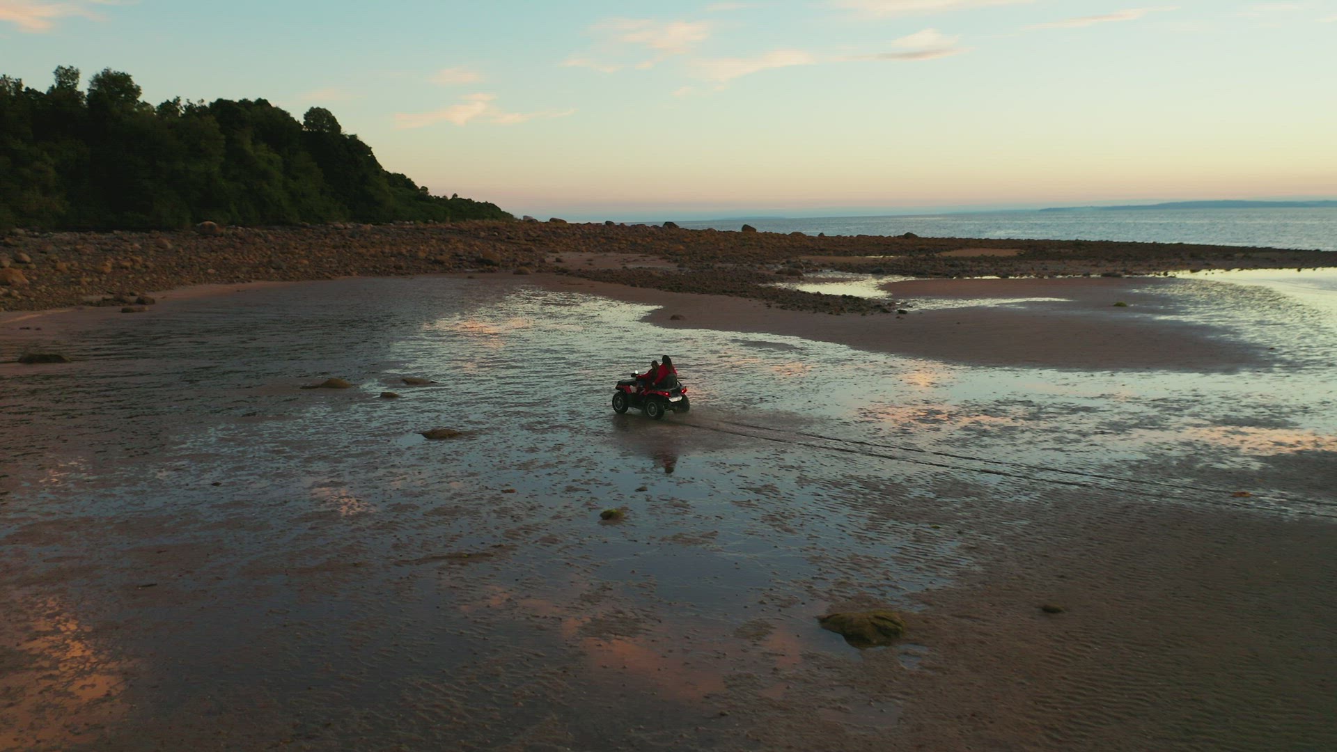 Aerial wide follow shot of a quad motor bike, riding over the wet sand of a coastal beach