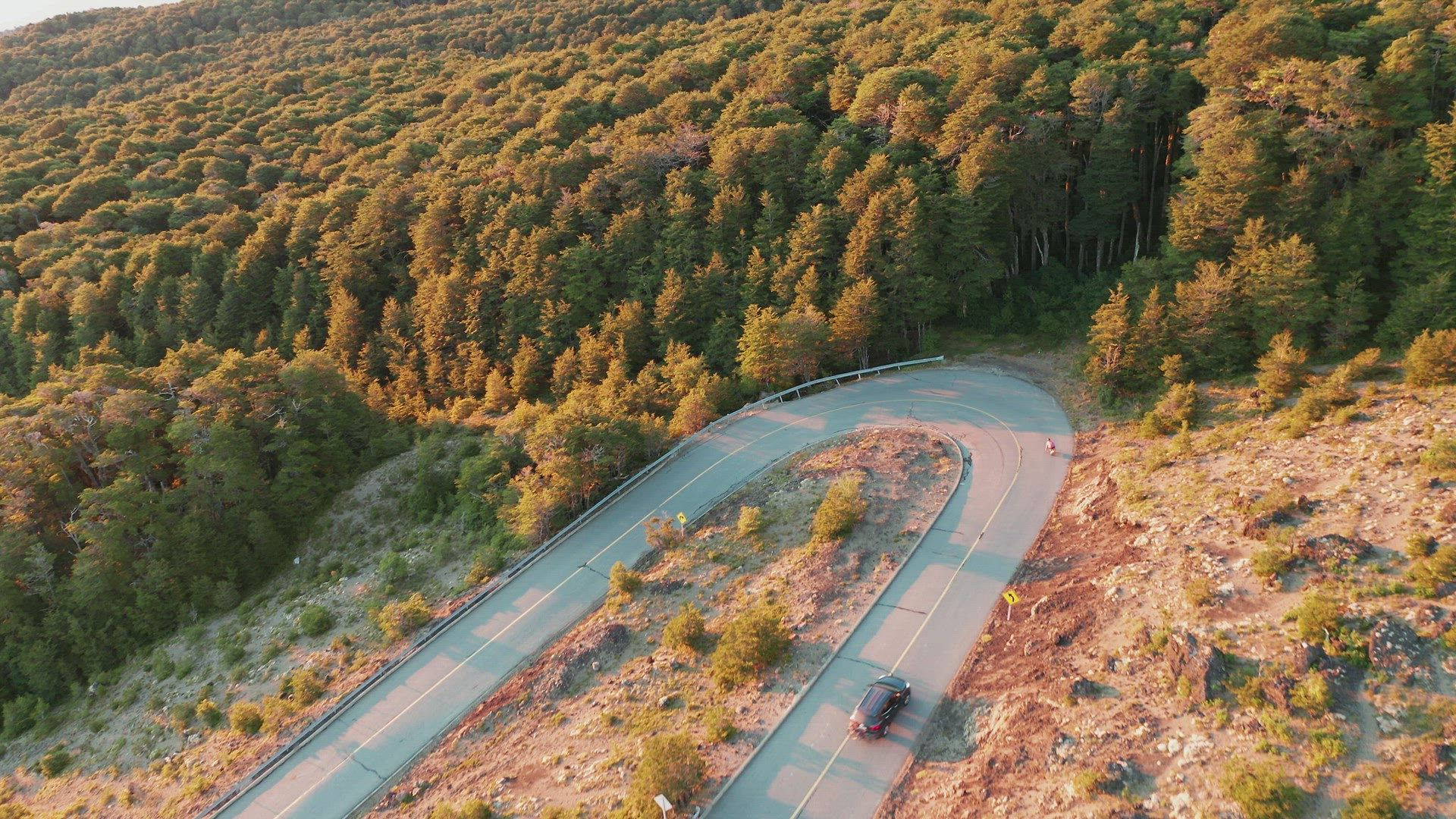 Aerial view of a longboard rider on a country road turning a sharp hairpin bend, bright sunlight.