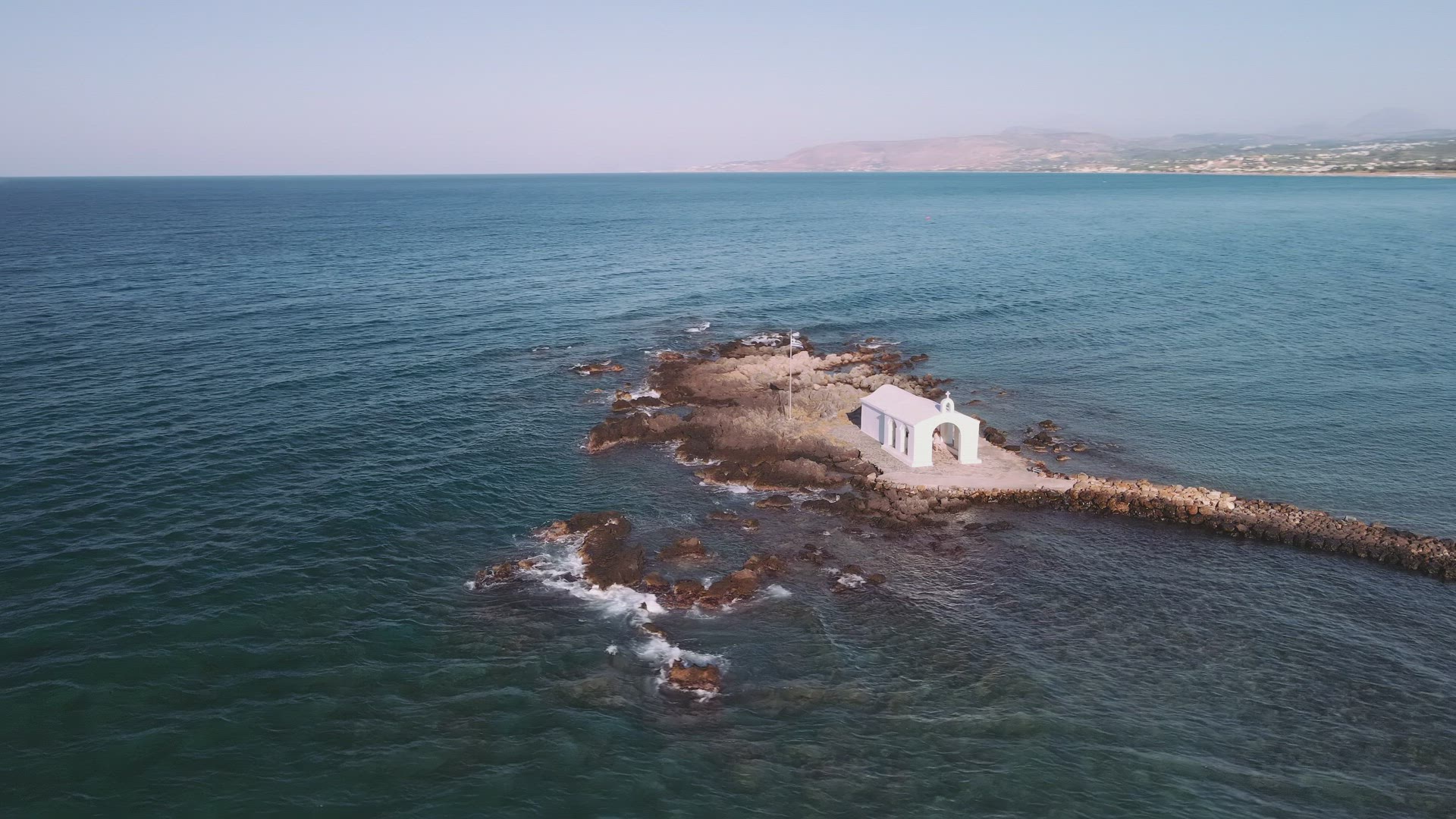 Aerial day view of whitewashed Greek Orthodox chapel around calm sea. 