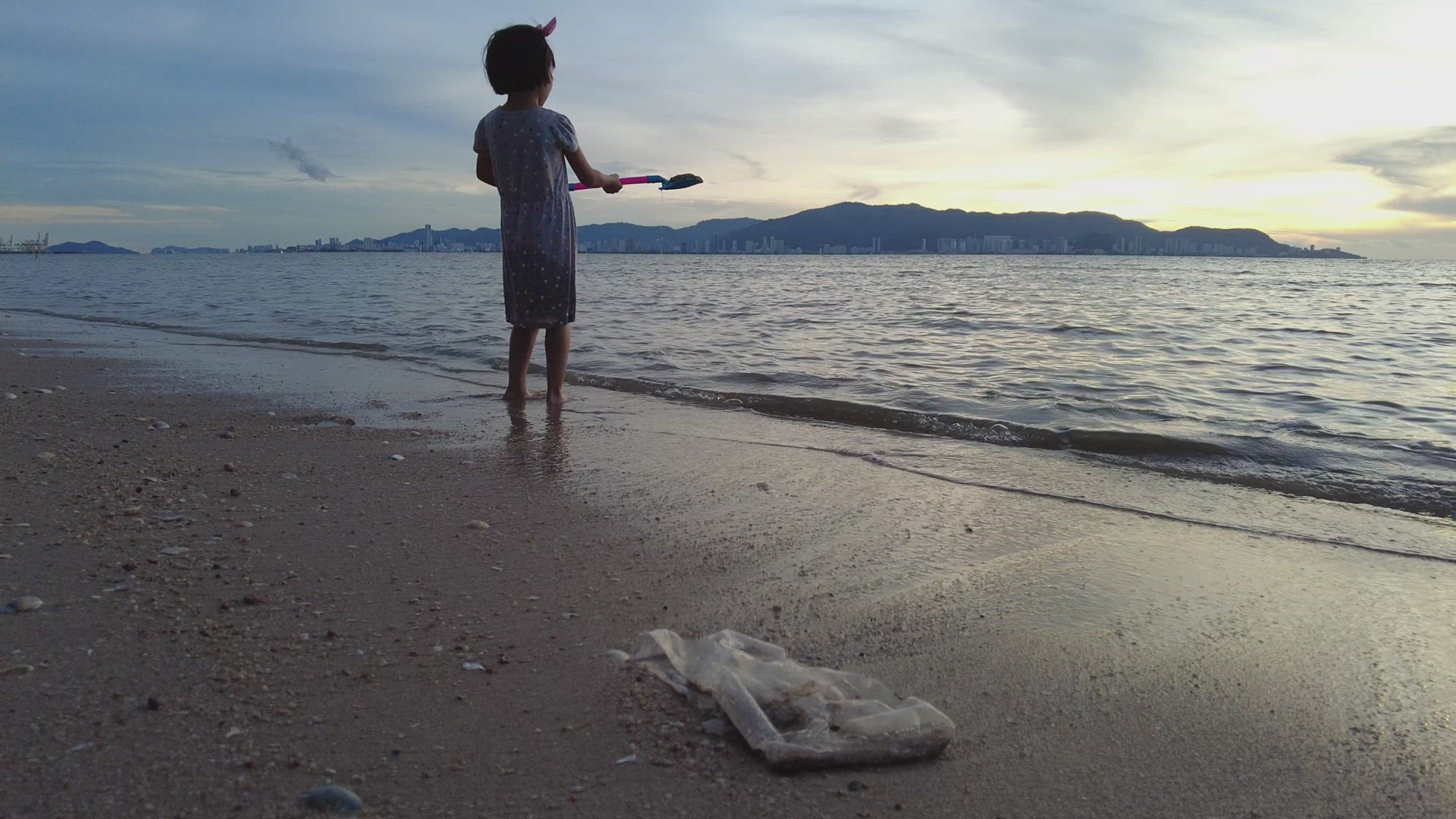 Little girl carry shovel with sand and play at sea near rubbish
