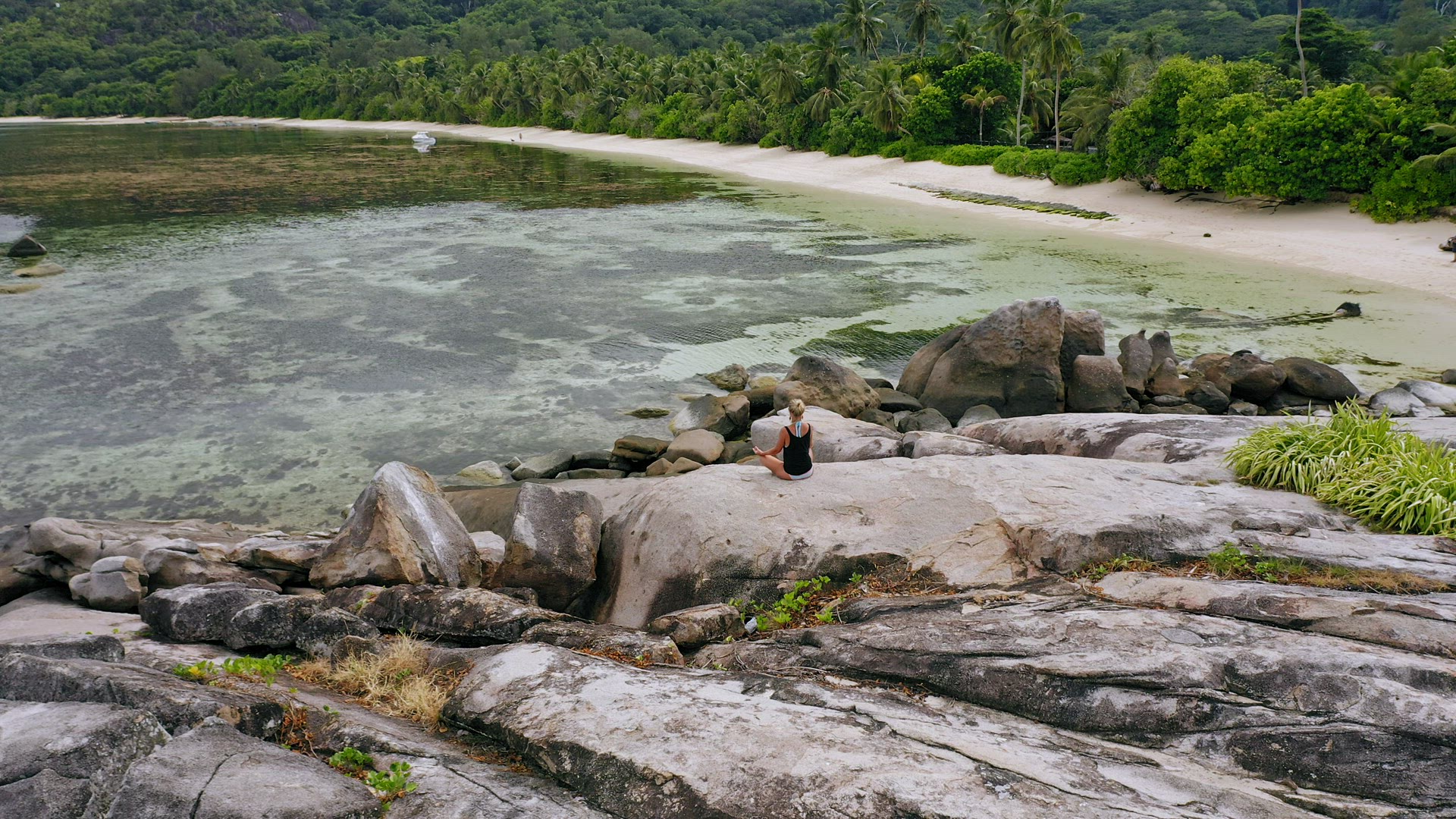 Aerial view of blonde tanned girl sitting in yoga pose and meditating on granite rock against exotic