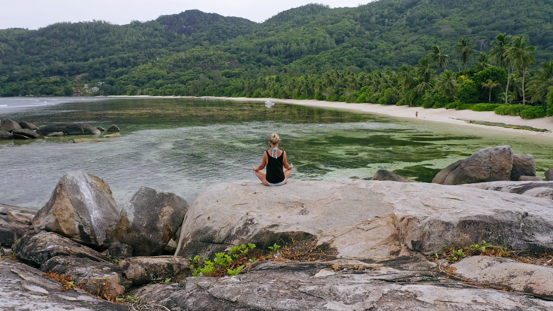 Aerial circle footage around blonde tanned girl sitting in yoga pose and meditating on granite rock 