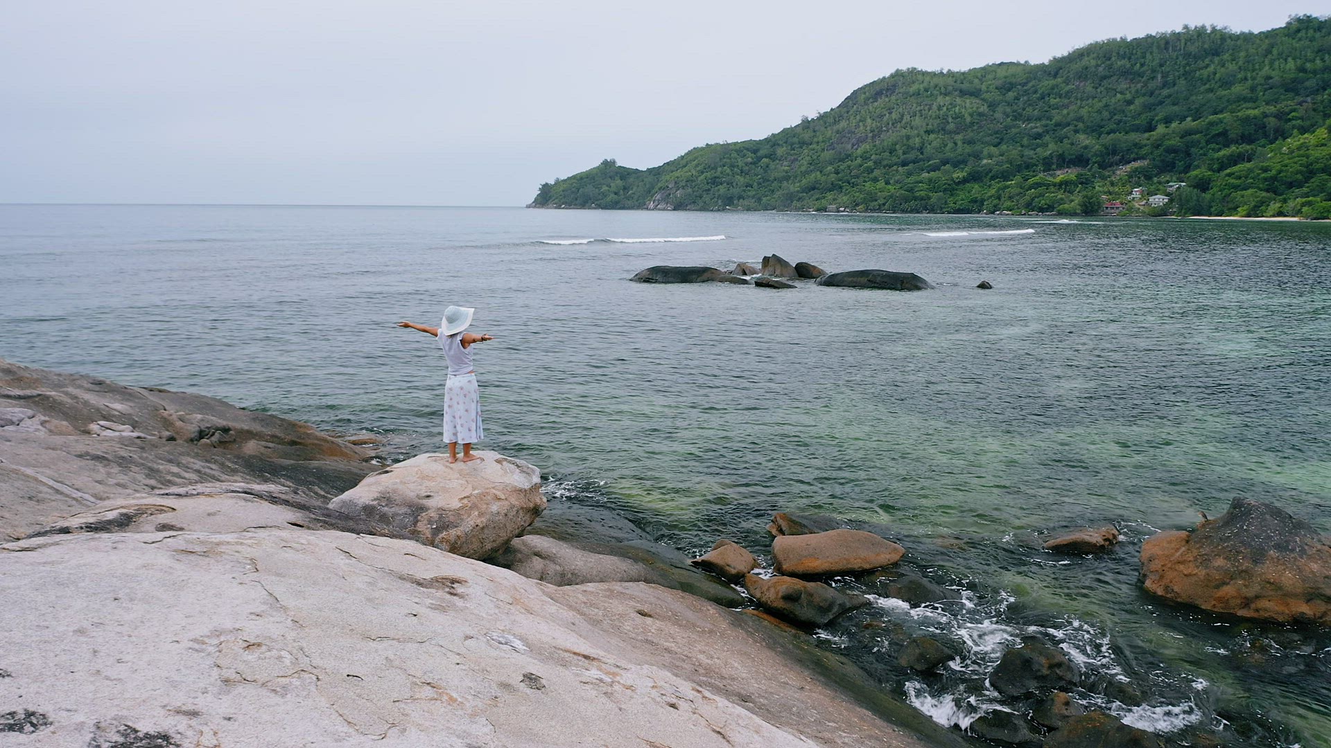 Seychelles, Mahe Island. Aerial circle footage of blonde girl with open hands staying on granite roc