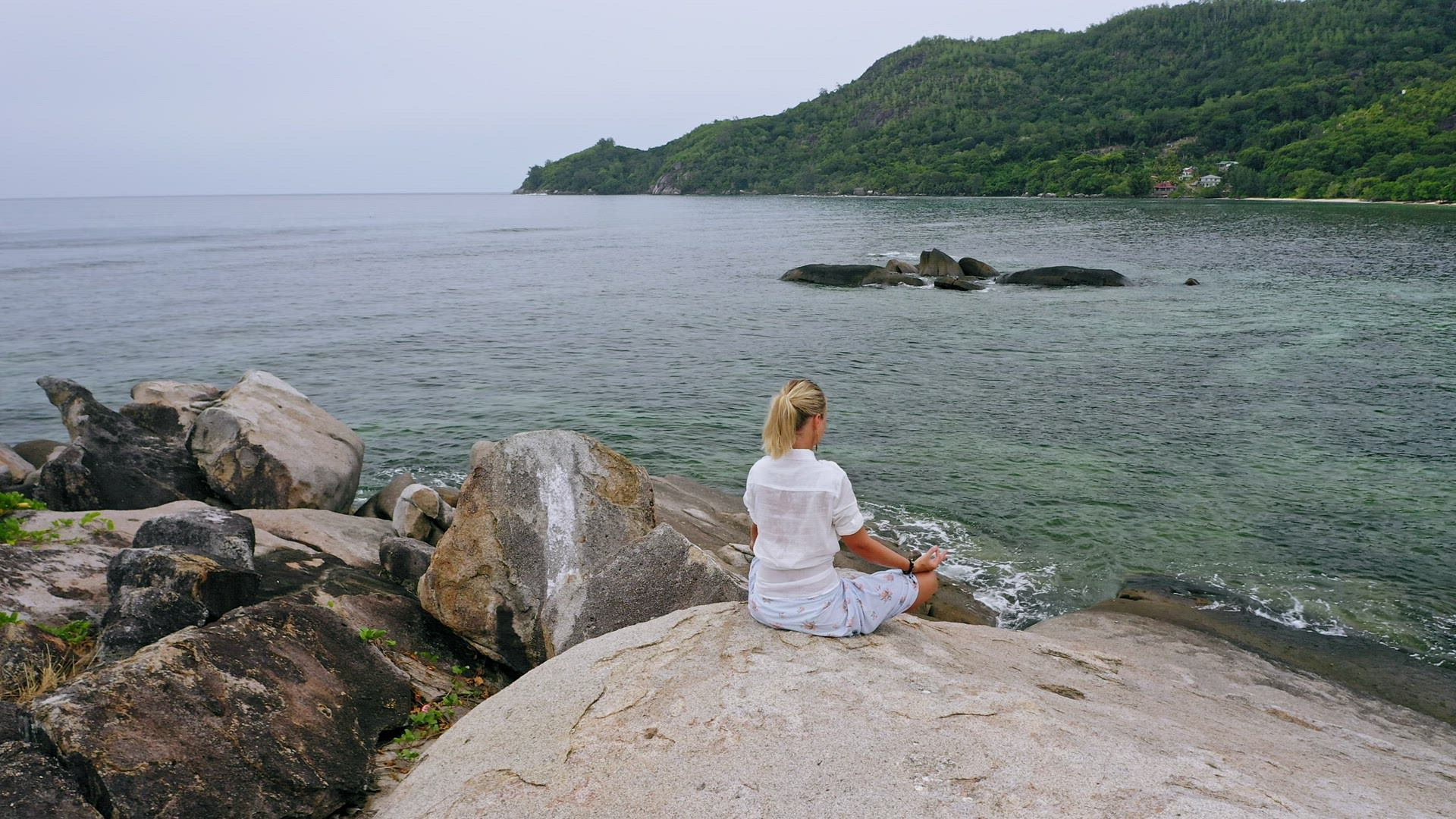 Aerial circling footage of blonde girl sitting in yoga pose on granite rock surrounded by exotic tro