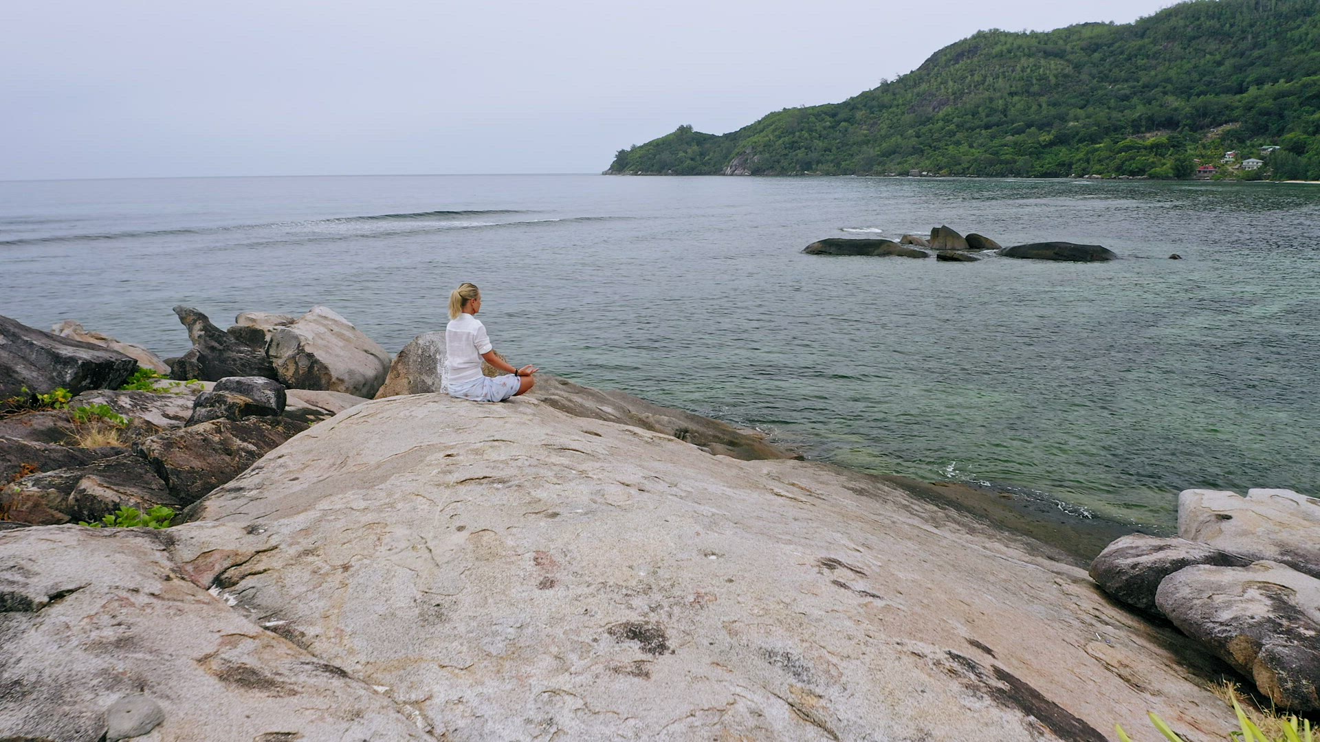 Aerial view of blonde tanned girl sitting in yoga pose and meditating on granite rock against exotic