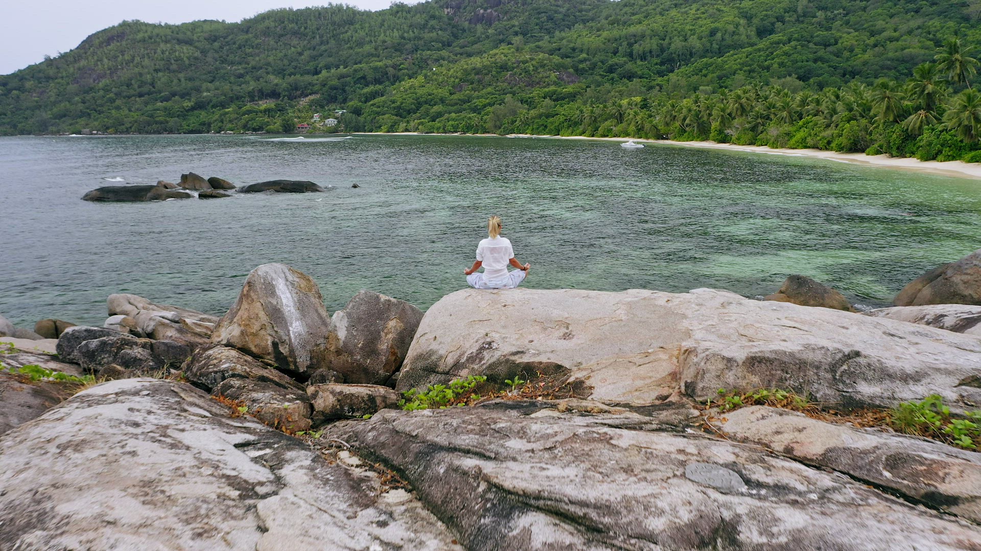 Aerial fly over blonde tanned girl sitting in yoga pose and meditating on granite rock against exoti