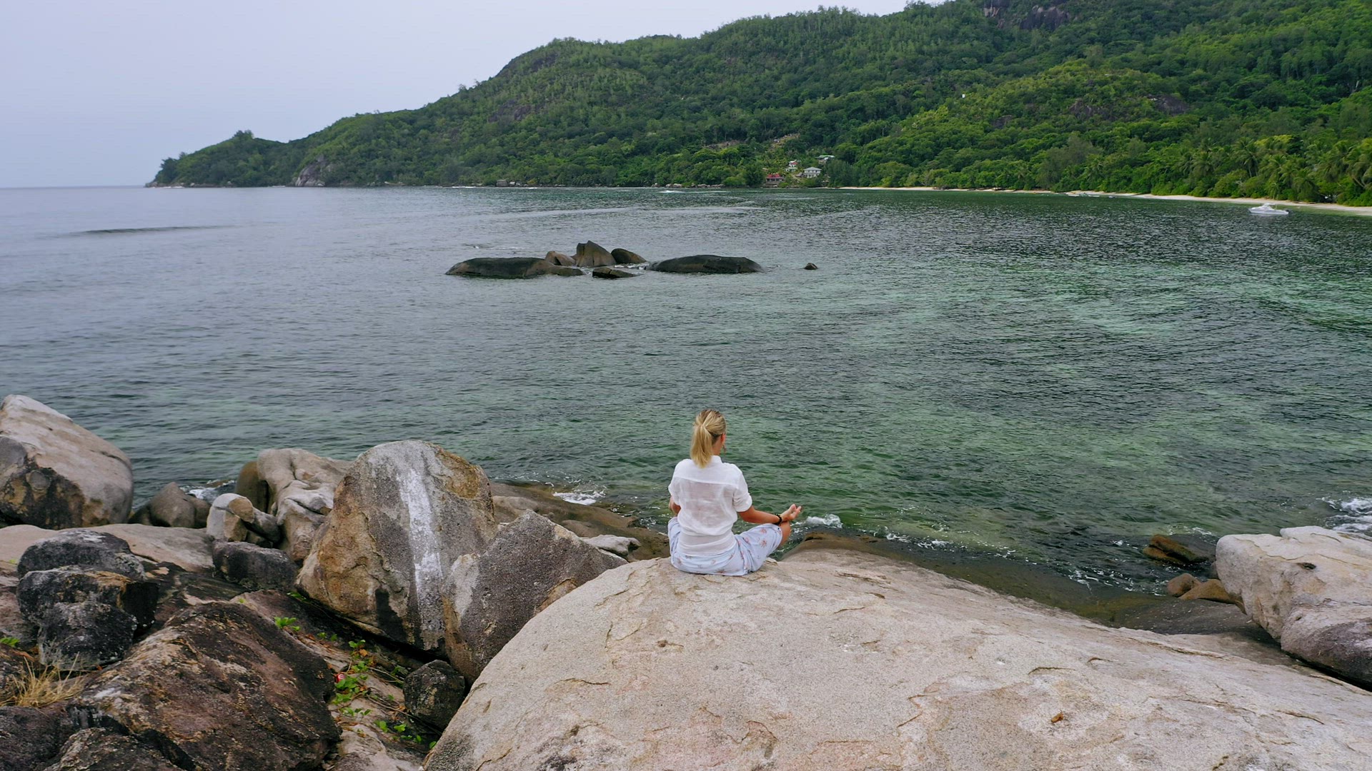 Aerial circle footage around blonde tanned girl sitting in yoga pose and meditating on granite rock 