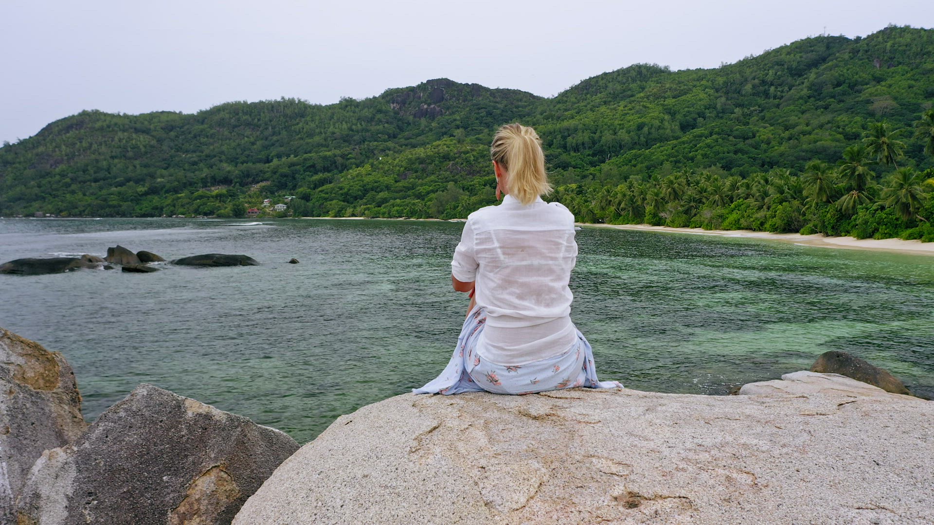 Aerial fly up reveal footage with blonde girl sitting on granite rock against exotic tropical lagoon
