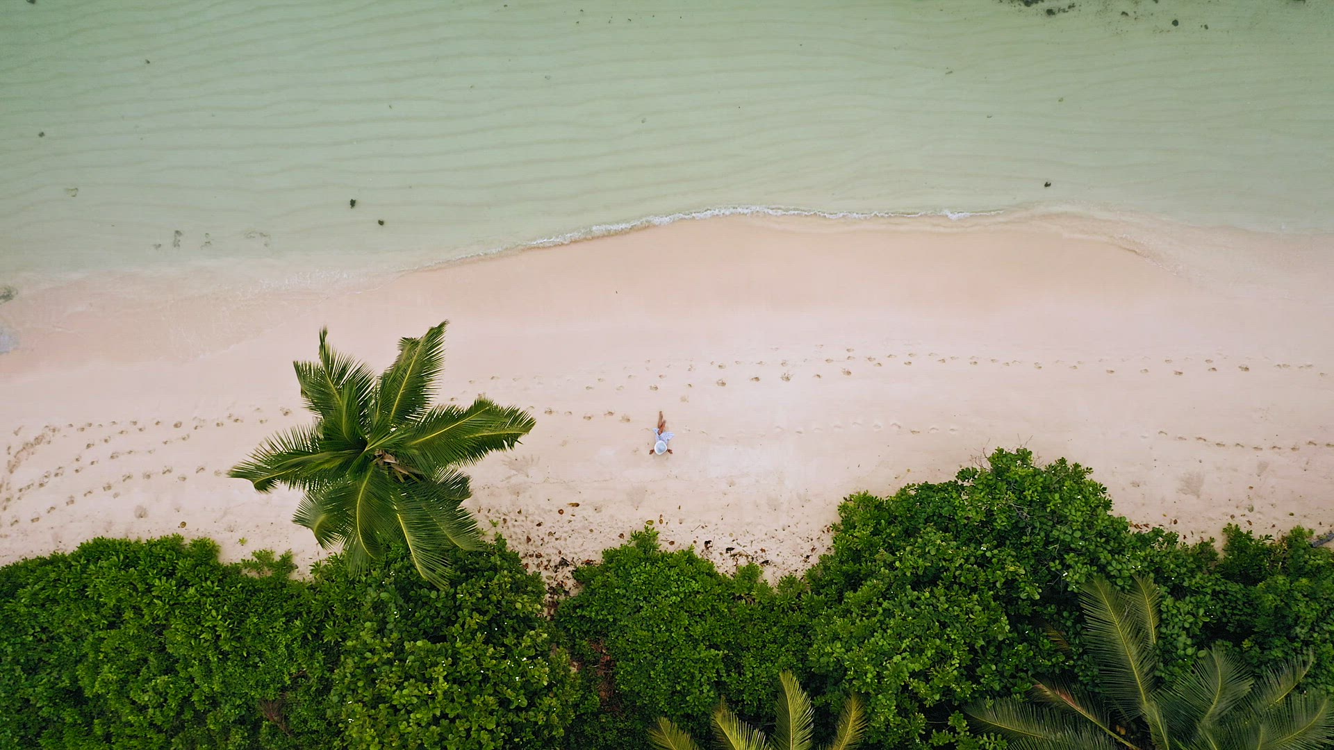Aerial vertical fly down footage. Girl sitting on tropical white sand beach surrounded by crystal cl