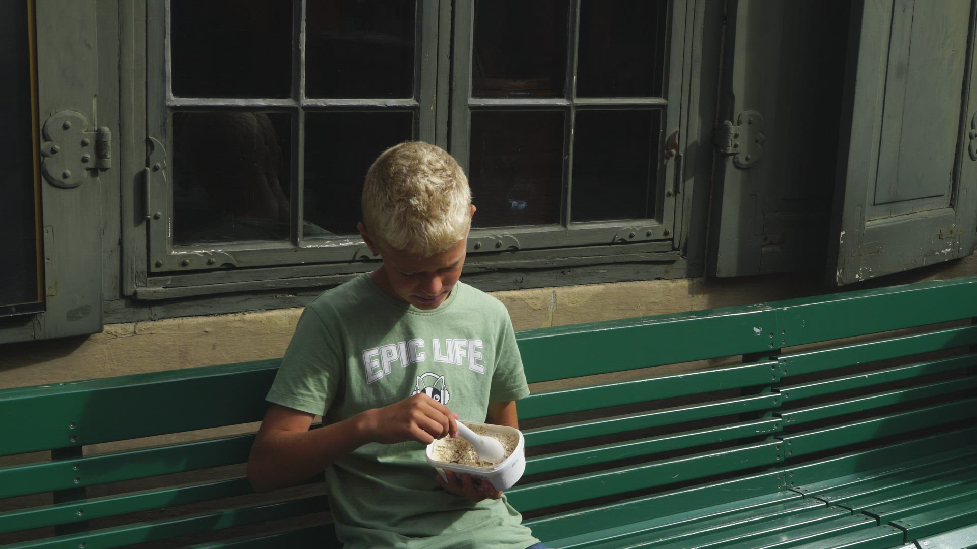 boy sitting on green bench eating icecream with spoon from plastic can
