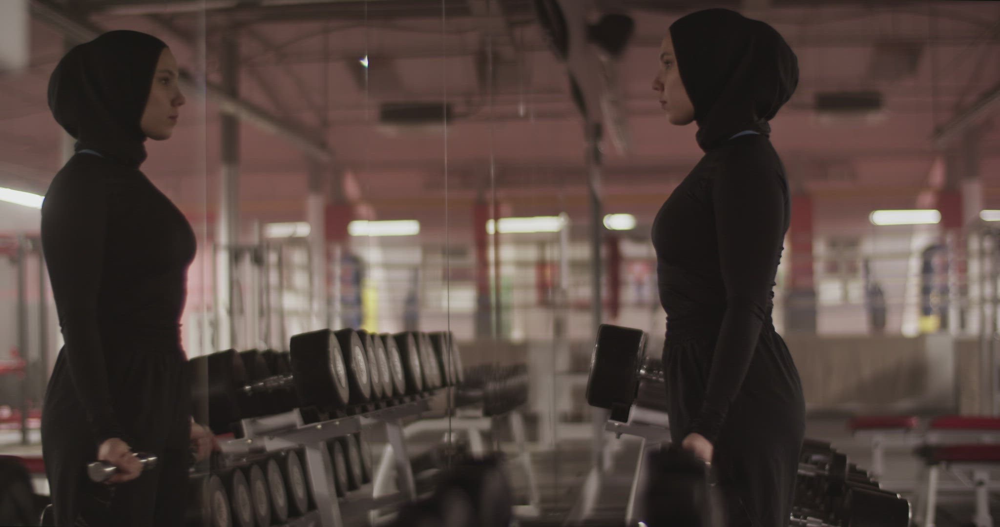 Muslim sportswoman doing biceps curls near mirror