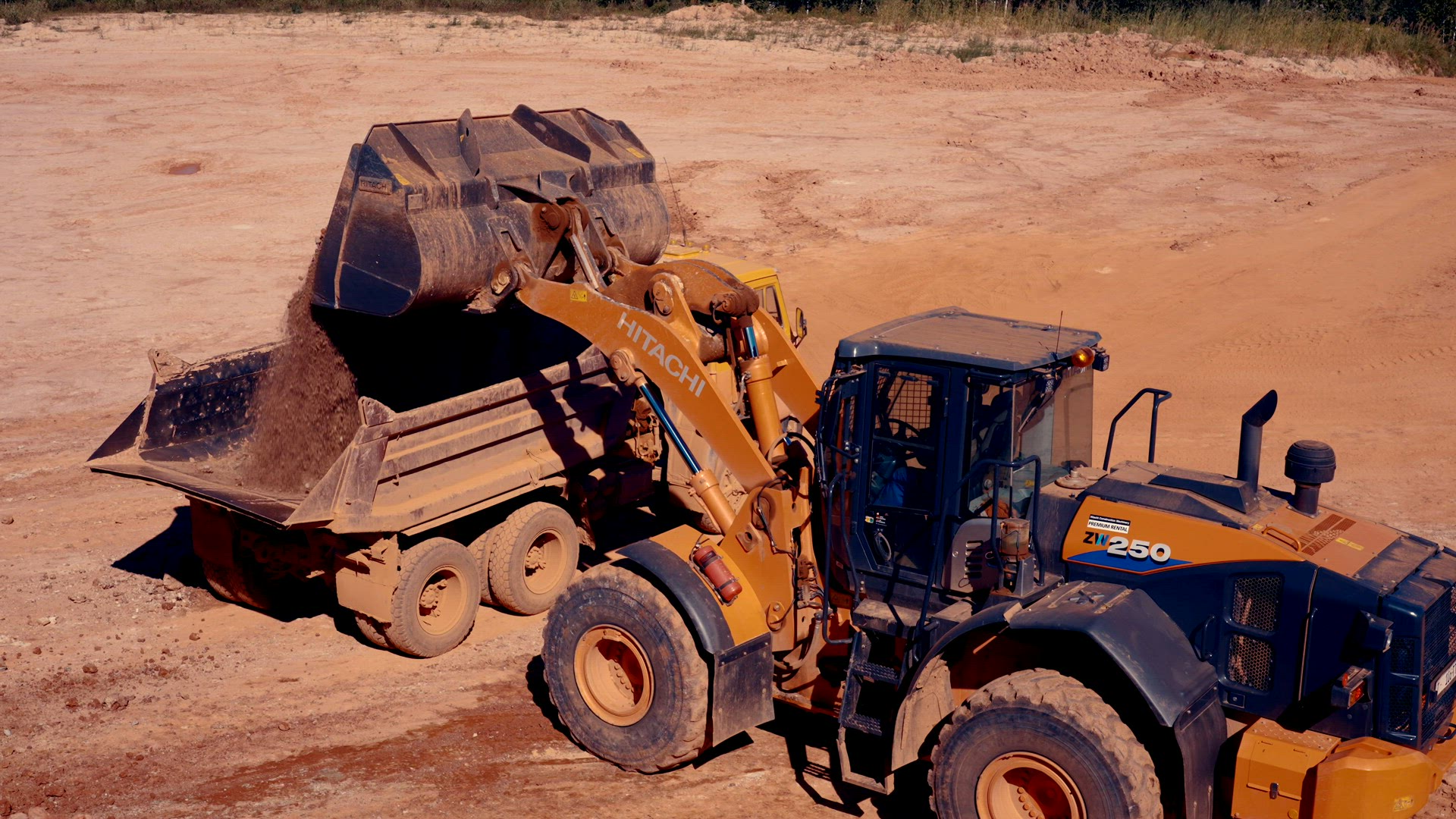 Loader empties its bucket filled with red clay into a dump truck.