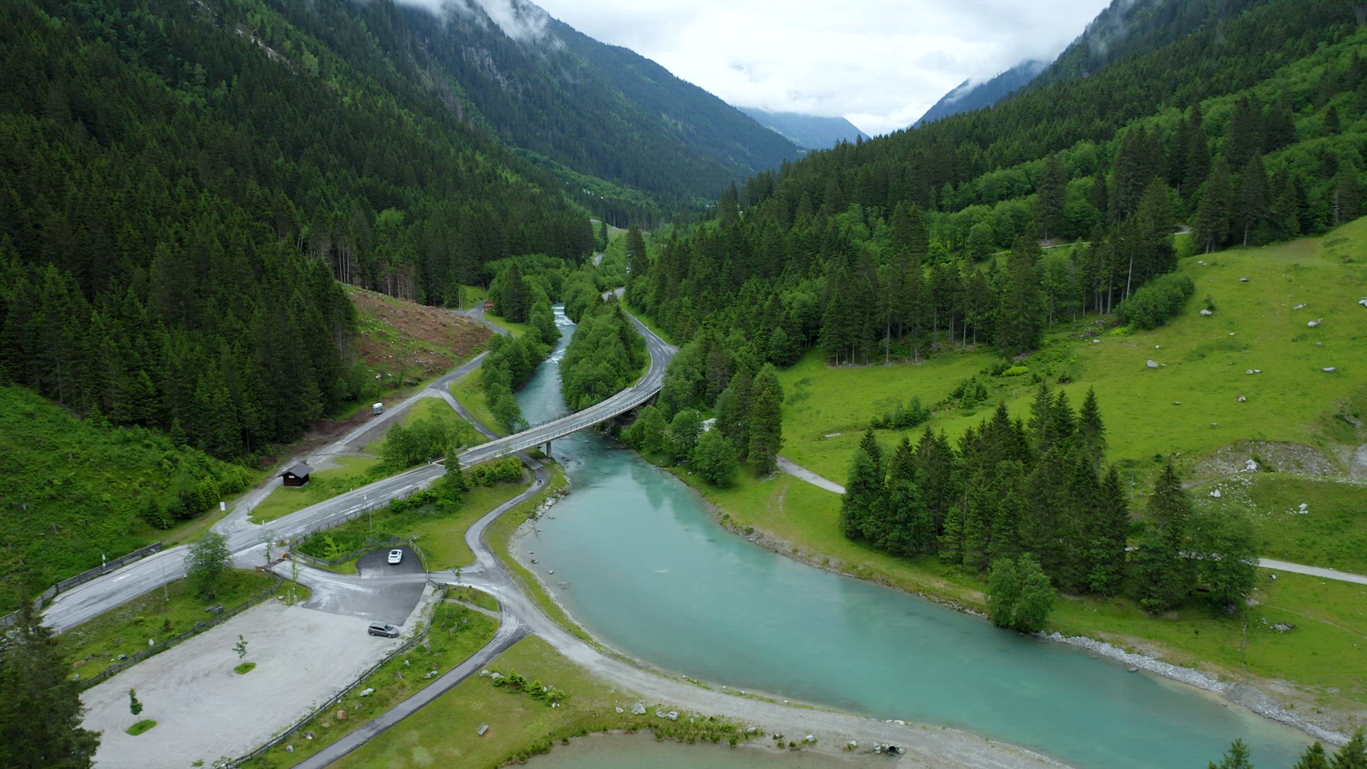 Aerial footage of mountain road crossing blue river in beautiful apline forest and waterfall in back