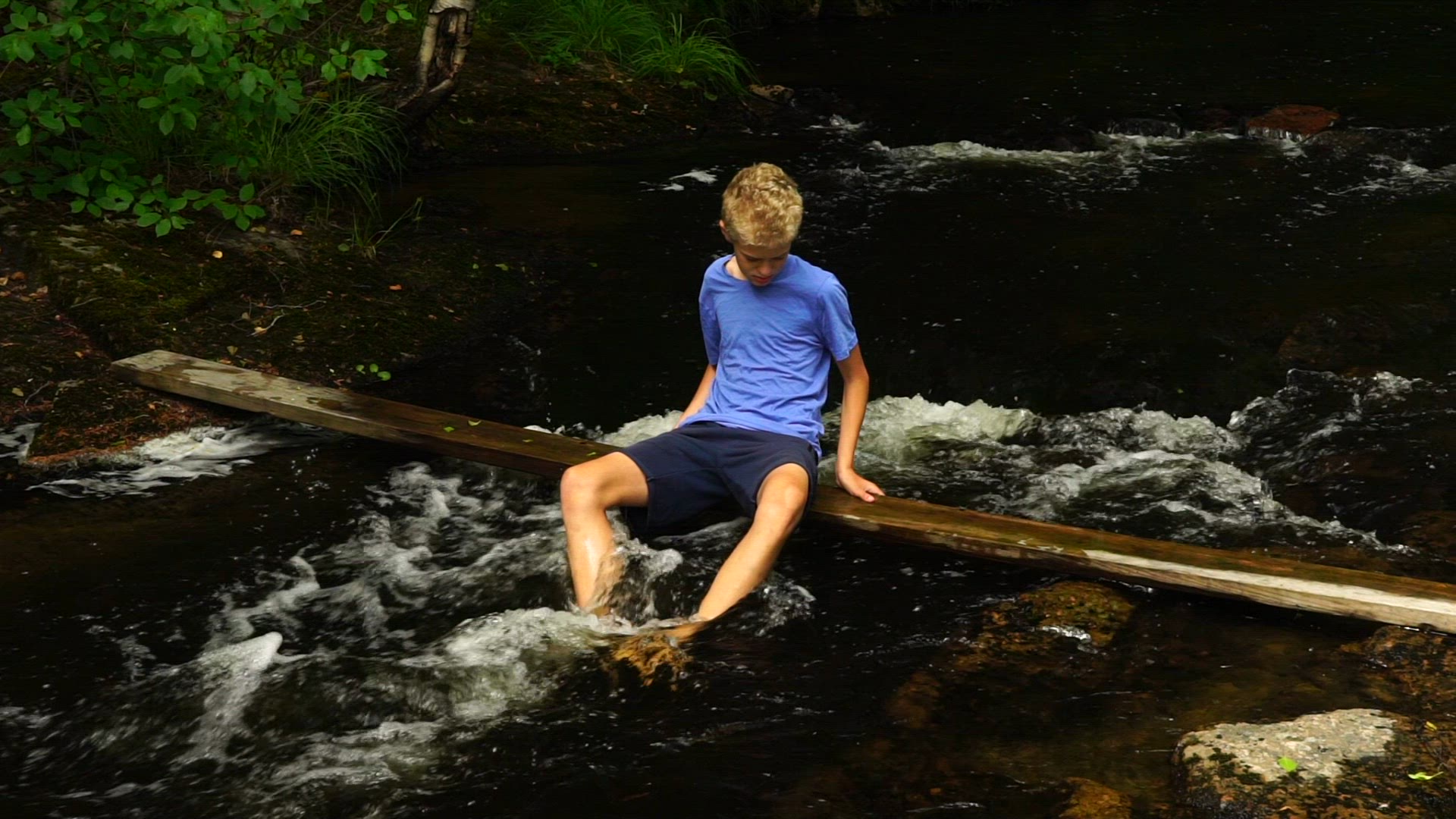 boy sitting on plank over river - feet in streaming water - rapids - slow motion shot