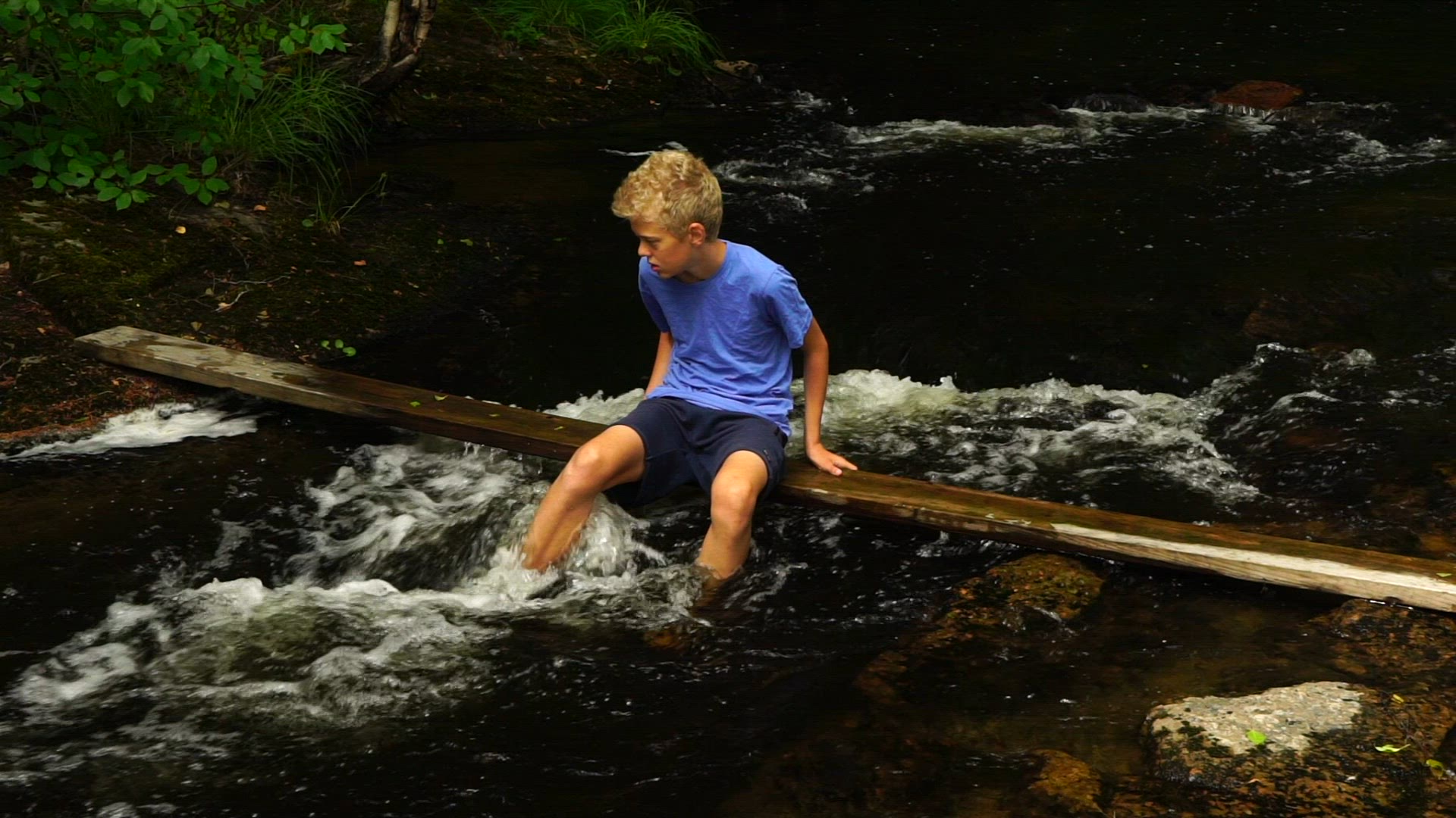 boy sitting on plank over river - feet in streaming water - rapids - slow motion shot