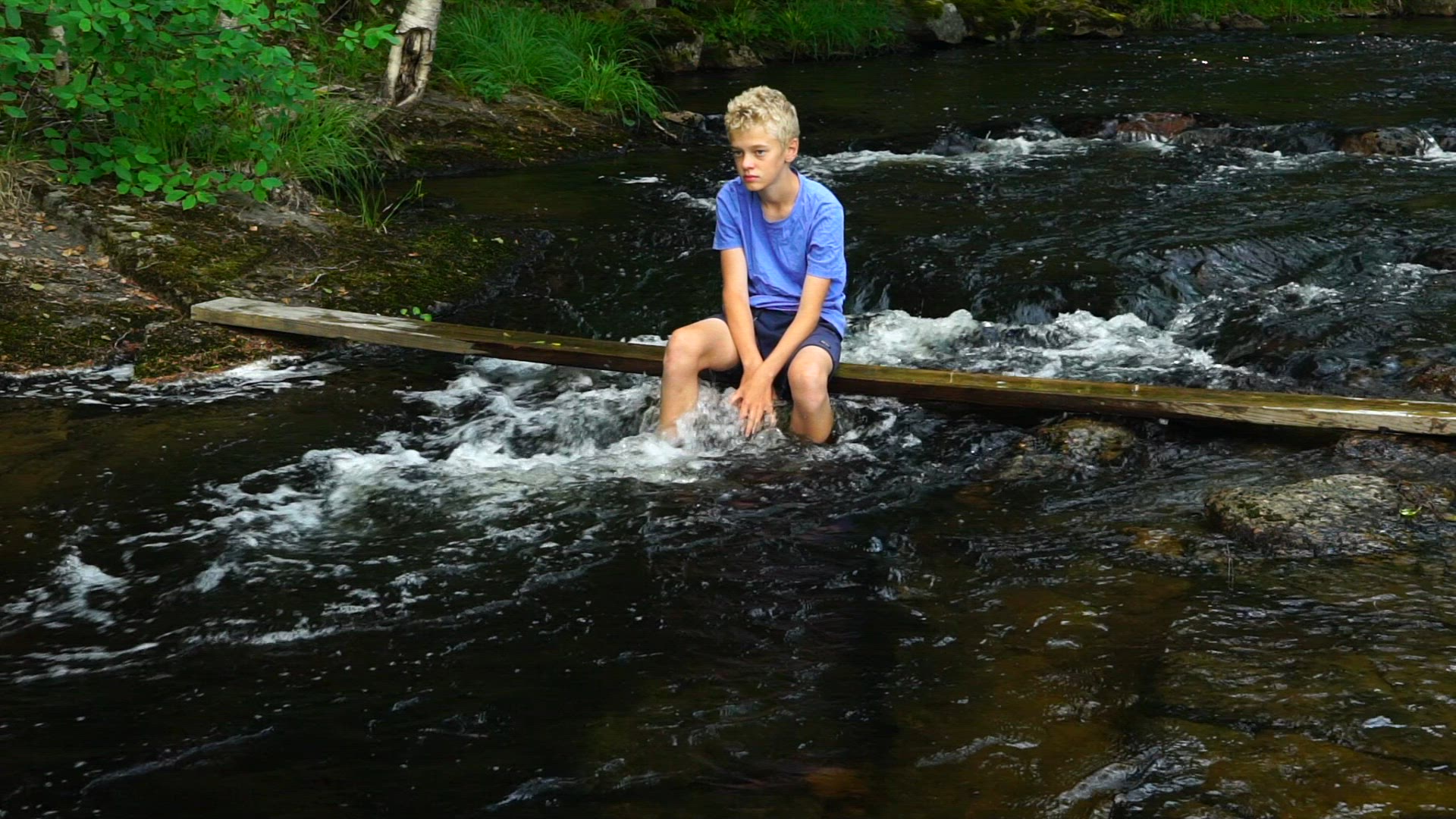 boy in blue t-shirt sitting on plank in river - water streaming under plank - slow motion shot