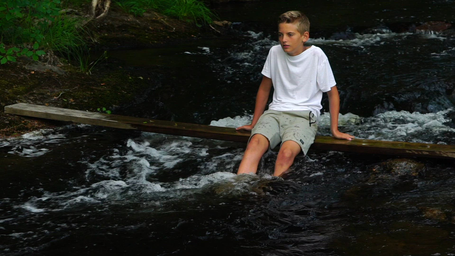 boy in white t-shirt sitting on plank i river - slow motion shot - rapids