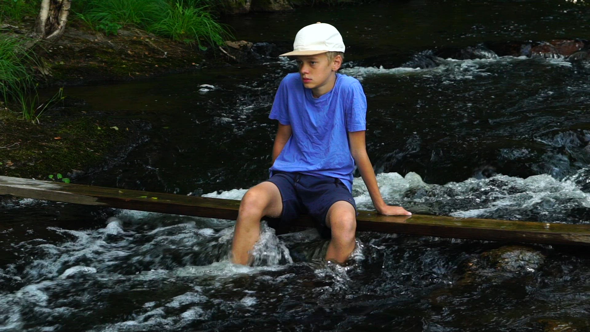 boy with cop sitting on plank in river - slow motion shot - streaming water in river