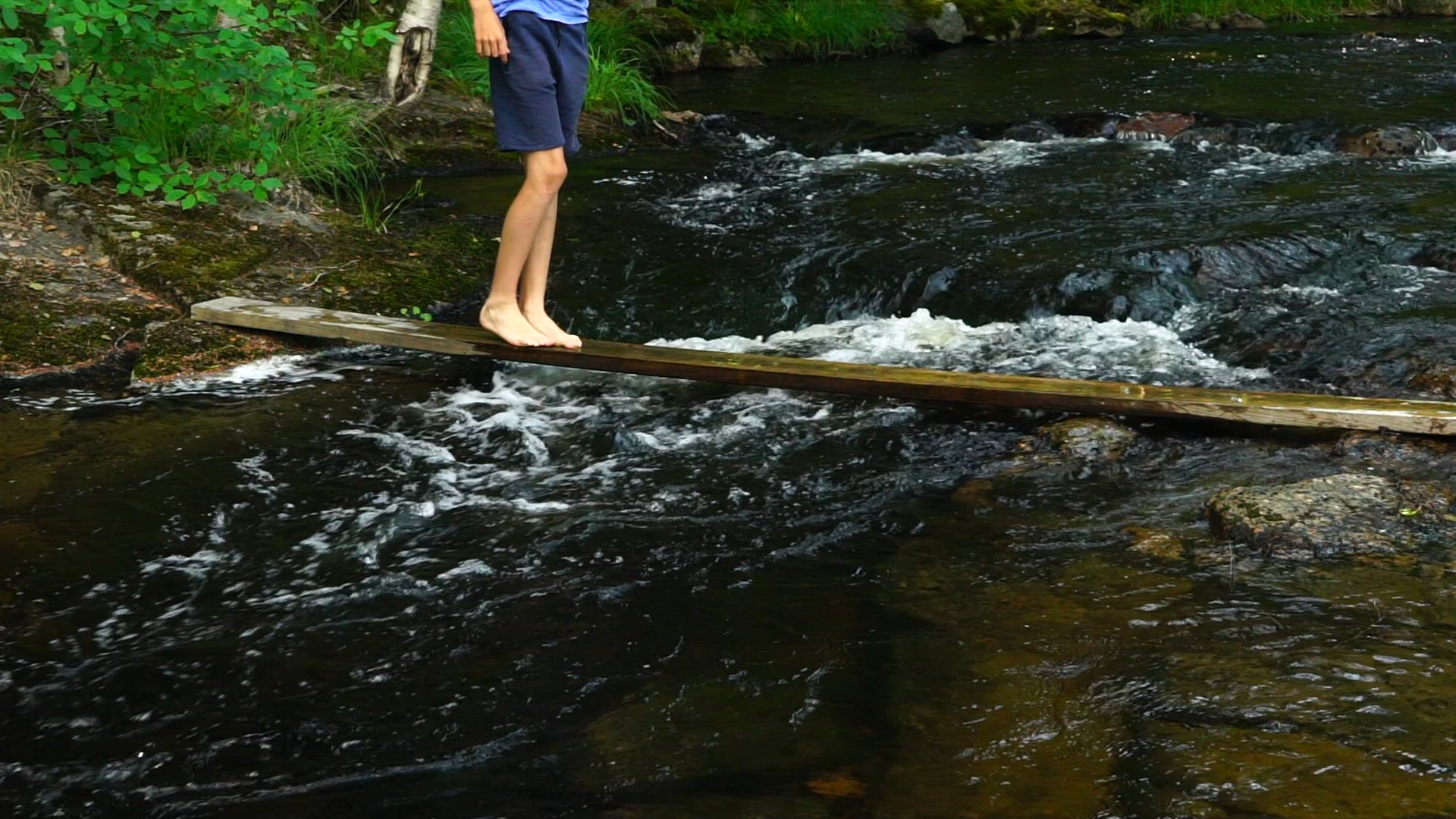 boy balancing on plank - walking over river with streaming water - close up feet and legs - slomo