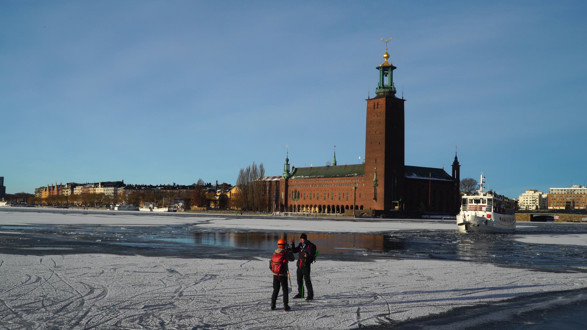 couple with skates standing on ice in central stockholm - city hall and white boat in background