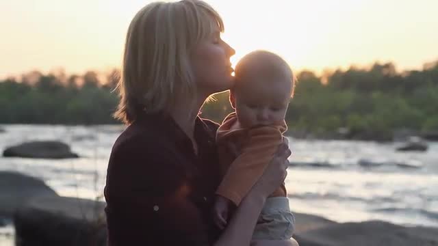Caucasian mother and baby hugging on riverbank at sunset