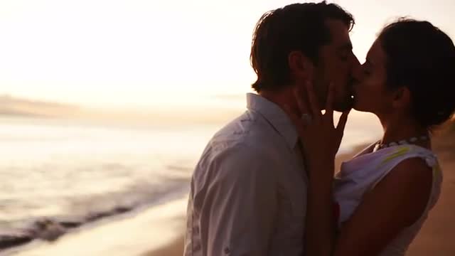 A silhouetted couple hold each other and kiss on the beach during magic hour