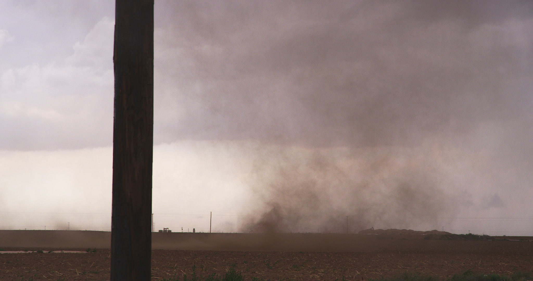 Small dusty tornado spins across red dirt field, past pump jack, Texas, 4K