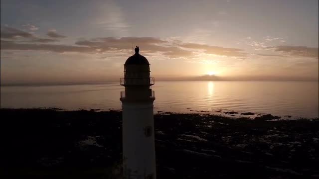 Aerial shot of a lighthouse during a beautiful sunrise in Scotland with coastline in background