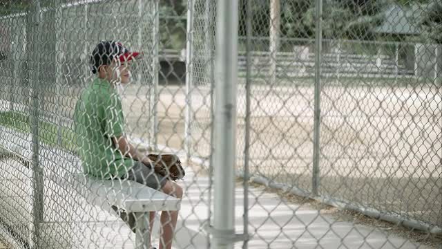 Slow motion push of boy sitting in dugout behind chain link fence.