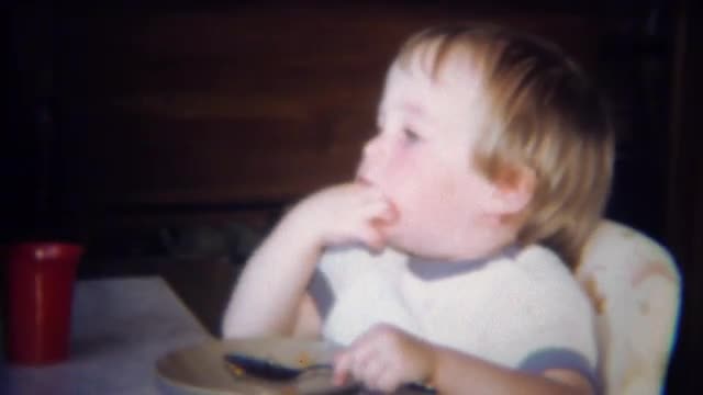 1971: Cute blonde boy eating pasta with hands and spoon in bowl.