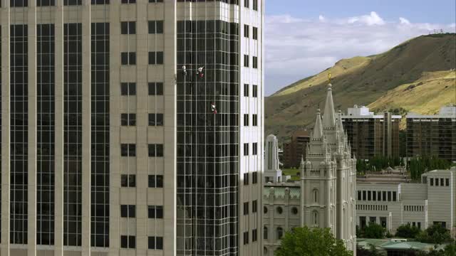 Three window washers on lines washing a highrise building. LDS Salt Lake Temple seen nearby.