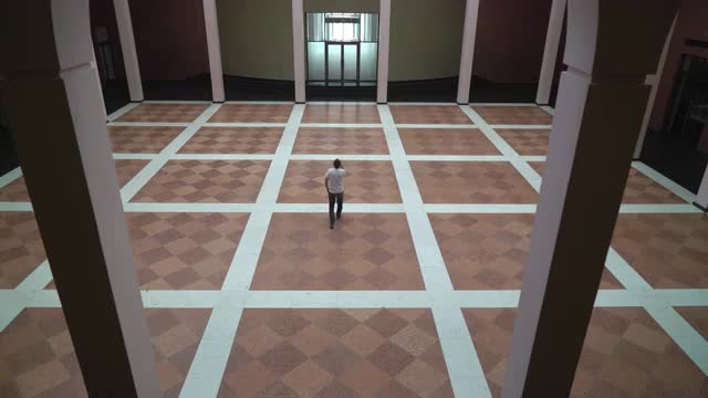 A man walks along in a large room with columns and talking on a cell phone.
