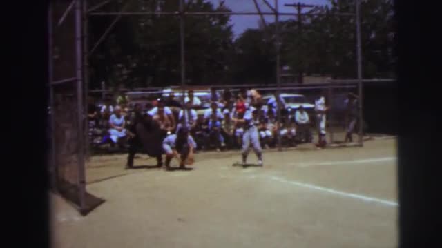 1972: an older boy in a gray baseball uniform and red batters cap is up to bat as spectators watch i