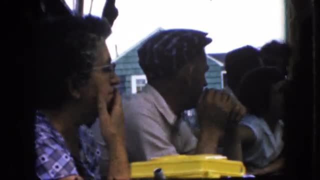 1969: group of people sitting at outdoor picnic table eating and talking with cape cod house in back