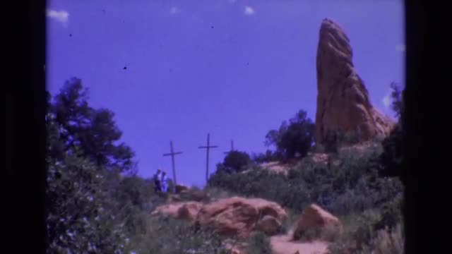 1967: two persons in blue are coming down from a rocky hill, crosses at the top of the hill. COLORAD