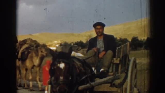 1962: a cigarette smoking fellow leads a caravan of camels with a horse drawn wagon cloudy sky IZMIR
