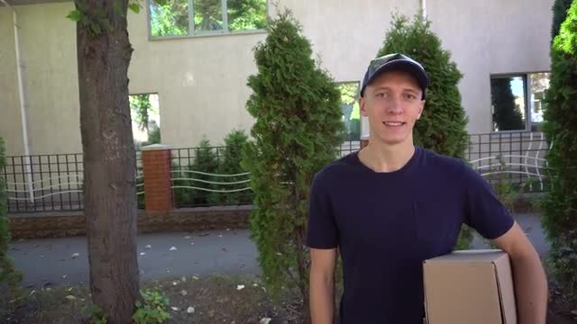 Pedestrian Courier Big Order in Hand and Smiling at Camera on Background of Trees and House, Stands 