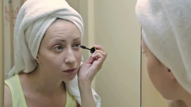 Beautiful young woman putting mascara in front of mirror in bathroom