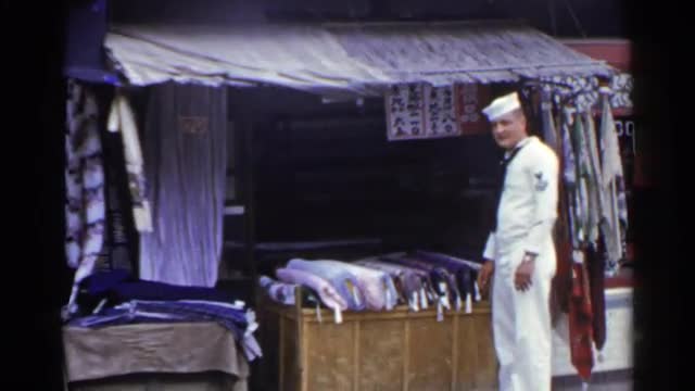 1953: sailor stands in front of outdoor stall displaying bolts of fabric JAPAN