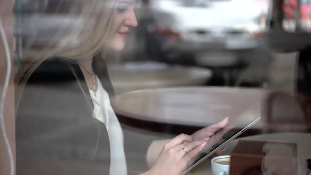 Young Business Woman Sits at a Table Drinking Coffee, Tea and Using Tablet Computer Typing on Tablet