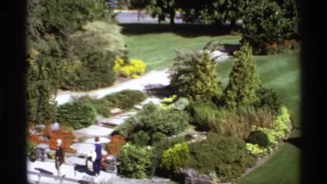 1974: a cute garden with different kind of plants and flowers two people are there to see the plants
