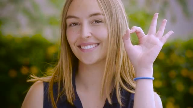 A smiling teenage girl with long blond hair gives an ok with her hand