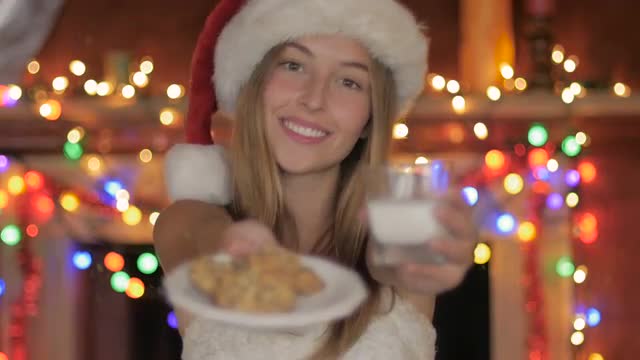 A beautiful young woman in santa hat offers cookies and milk at Christmas