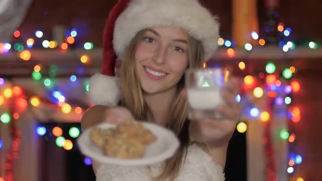 A beautiful young woman wearing a Santa hat offers cookies and milk