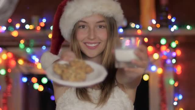 A beautiful woman in a santa hat offers cookies and milk with rack focus