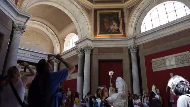 Footage tilts down from a ceiling fresco in the Vatican Museums to tourists taking photos.